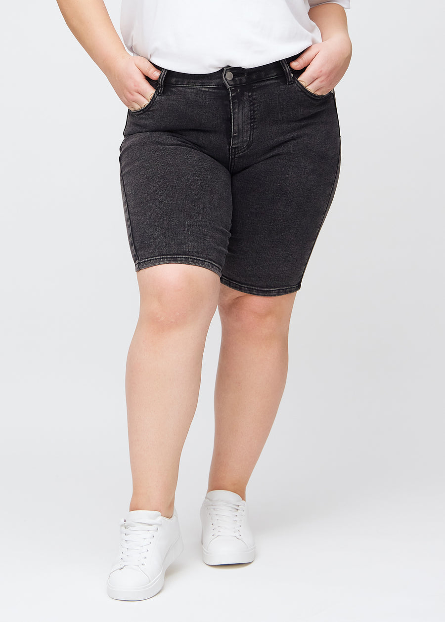 Mörkgrå denim skinny middle shorts, modellnamn Shadows, som sitter tätt över låret och slutar precis ovanför knät på en plus size-modell, sedd framifrån.