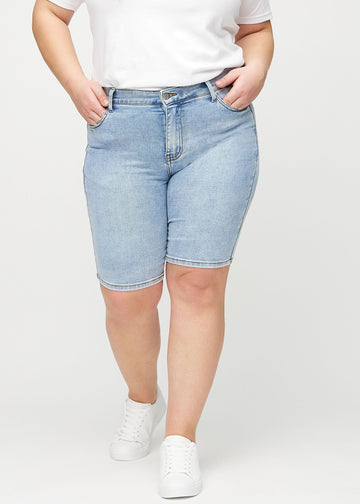 Ljus denim skinny middle shorts, modellnamn Waves, som sitter tätt över låret och slutar precis ovanför knät på en plus-size-modell, sett framifrån.