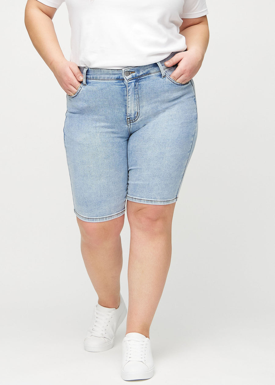 Ljus denim skinny middle shorts, modellnamn Waves, som sitter tätt över låret och slutar precis ovanför knät på en plus-size-modell, sett framifrån.