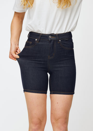 Modell sträcker mörkblå denimshorts för dam i färgen Midnights vid låret för att visa materialets stretch.