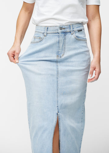 Model strækker den lange, lyseblå denimnederdel i farven Waves ved hoften for at vise materialets stretch.