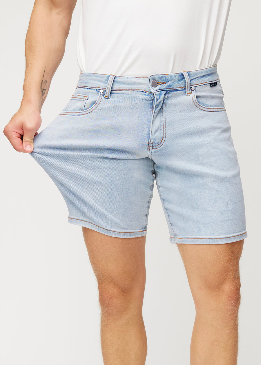 Model strækker lyseblå denim shorts til mænd i farven Waves ved låret for at vise materialets stretch.