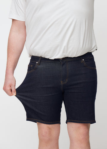 Plus-size modell sträcker mörkblå denimshorts för herr i färgen Midnights vid låret för att visa stretch.