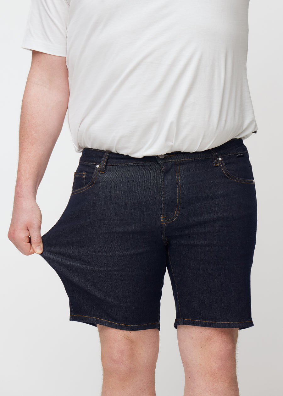 Plus-size modell sträcker mörkblå denimshorts för herr i färgen Midnights vid låret för att visa stretch.